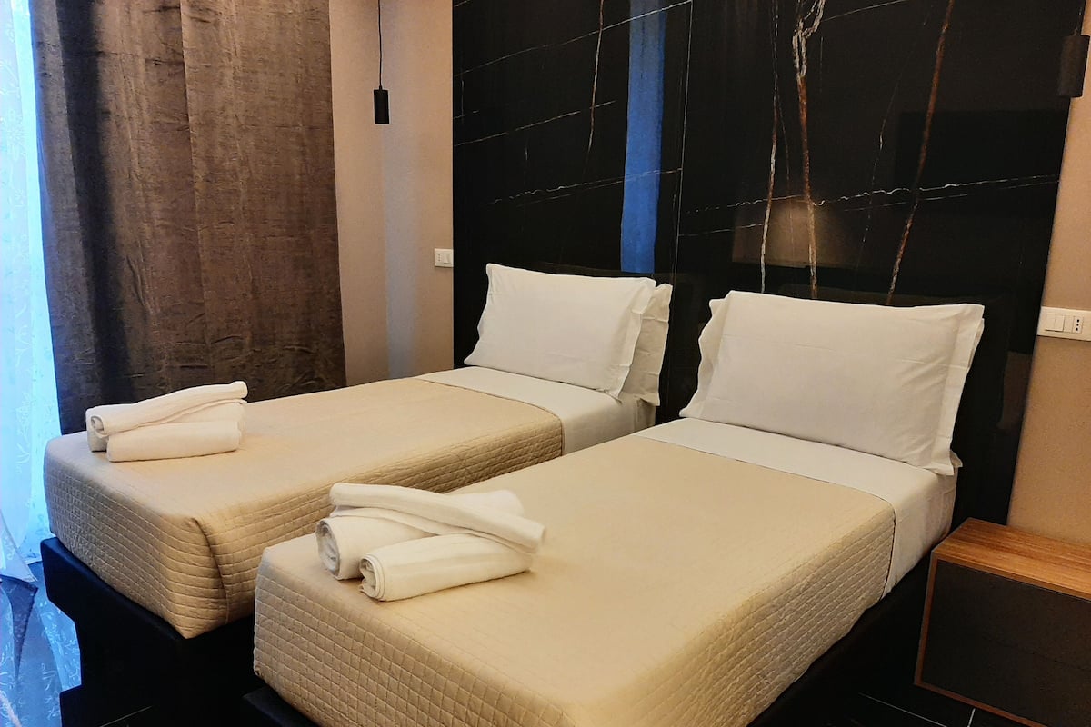 A hotel room with two single beds, each with a quilted beige bedspread and white pillows. There is a wooden nightstand with a black surface and a white power outlet on the wall.