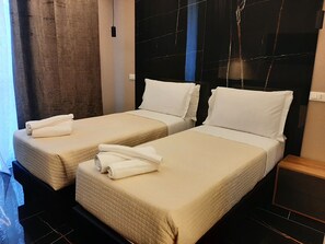 Une chambre d’hôtel avec deux lits simples, chacun recouvert d’une courtepointe beige matelassée et agrémenté d’oreillers blancs. On y trouve une table de chevet en bois, avec un plateau noir et une prise électrique blanche fixée au mur.