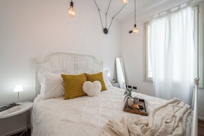 Un lit bien fait, avec une literie blanche et des coussins décoratifs, une table de chevet avec une lampe, et un plateau avec des biscuits et une tasse.