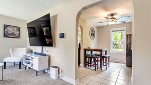 Sala de estar com TV de tela plana, móvel de entretenimento branco e poltrona branca. Há uma área de jantar com mesa e cadeiras de madeira.