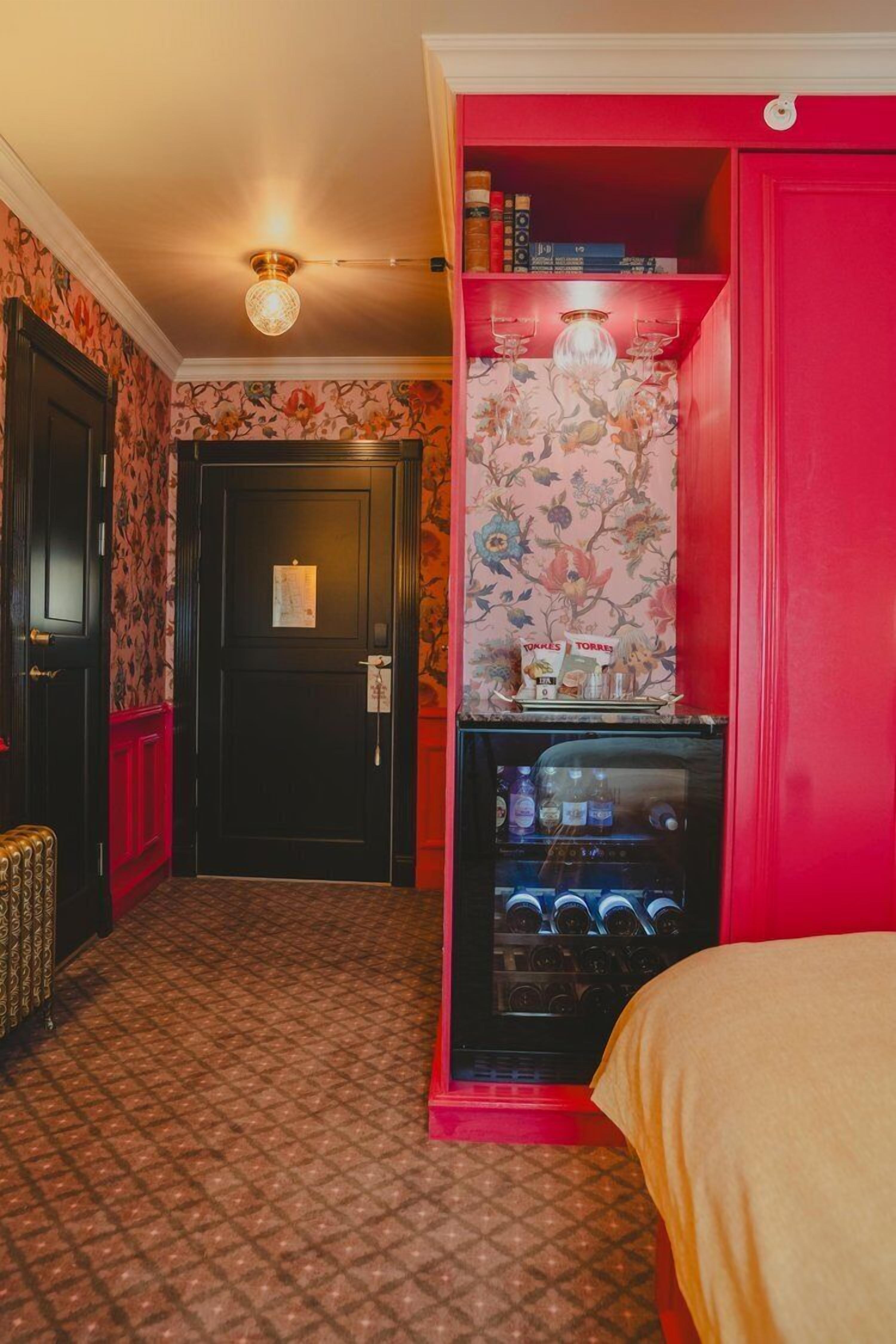 A hotel room with a patterned floor, a minibar, a red wall, and a door with a notice on it.