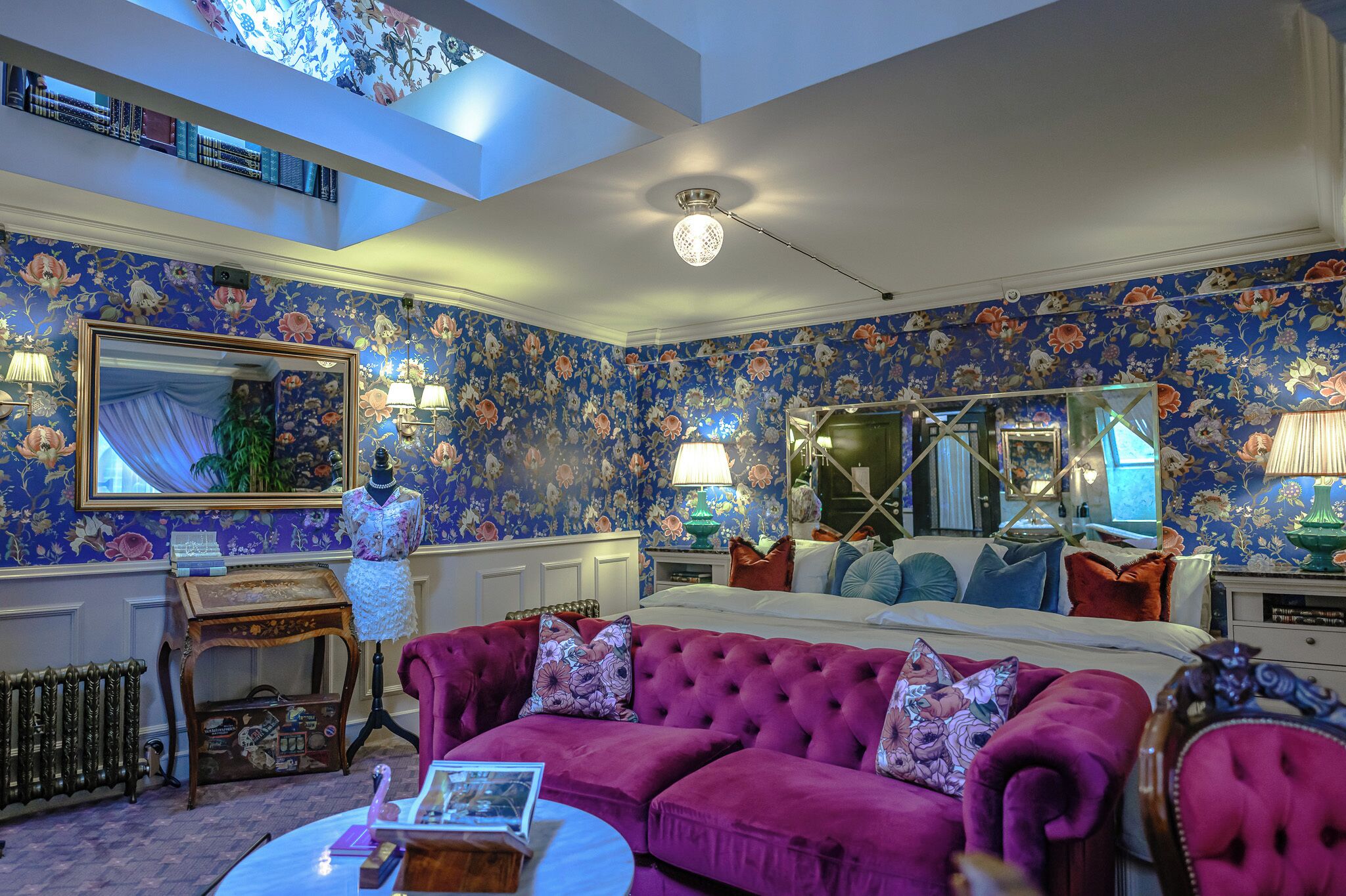 A room with a floral wallpaper, a large mirror, a tufted sofa, and a ceiling fan.