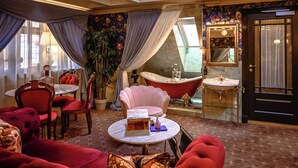 A room with a red sofa, a round table with a book and a vase, a pink armchair, a bathtub, a mirror, and a door with a curtain. - Charmante Skostredet Hôtel (Bergen)