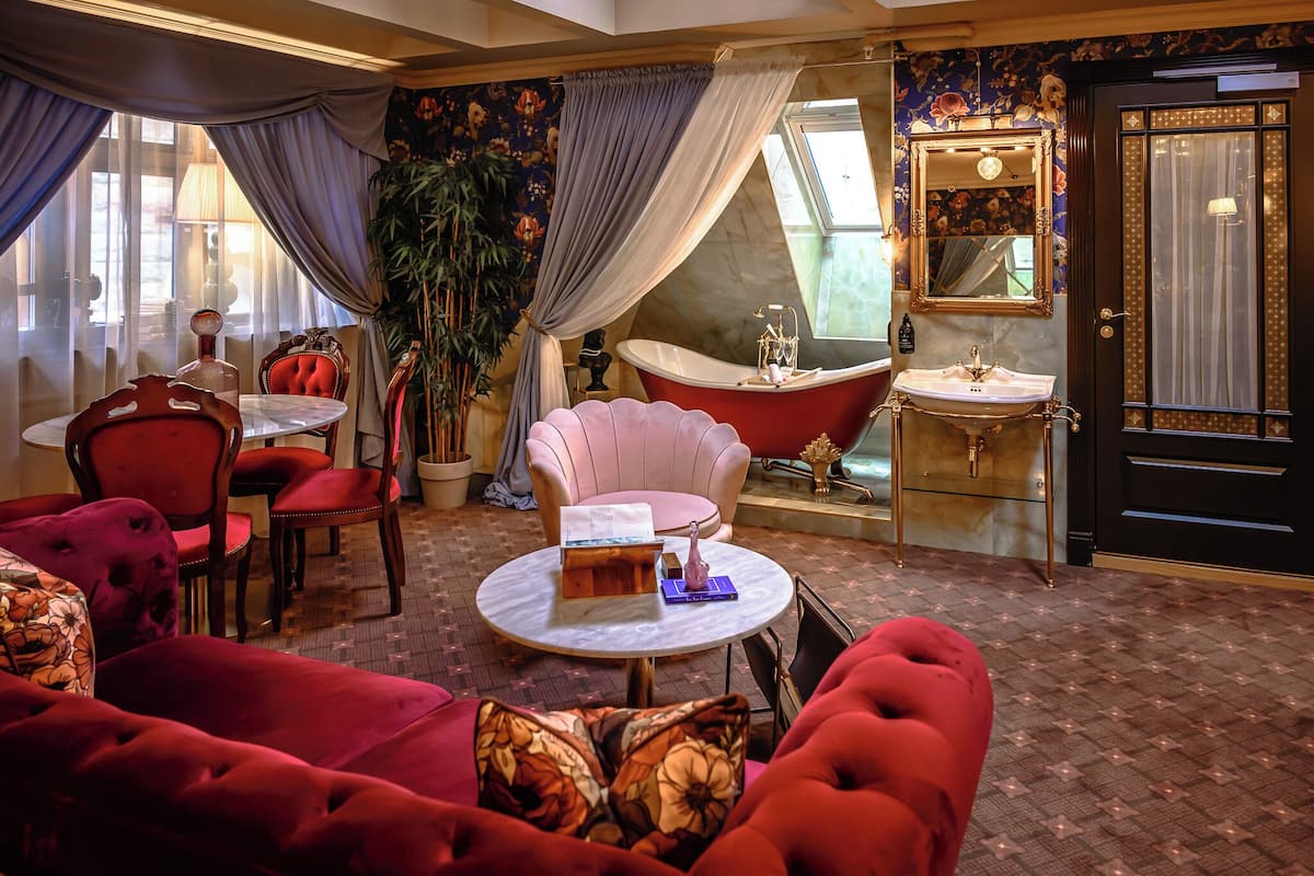 Ein Zimmer mit einem roten Sofa, einem runden Tisch mit einem Buch und einer Vase, einem pinken Sessel, einer Badewanne, einem Spiegel und einer Tür mit Vorhang.