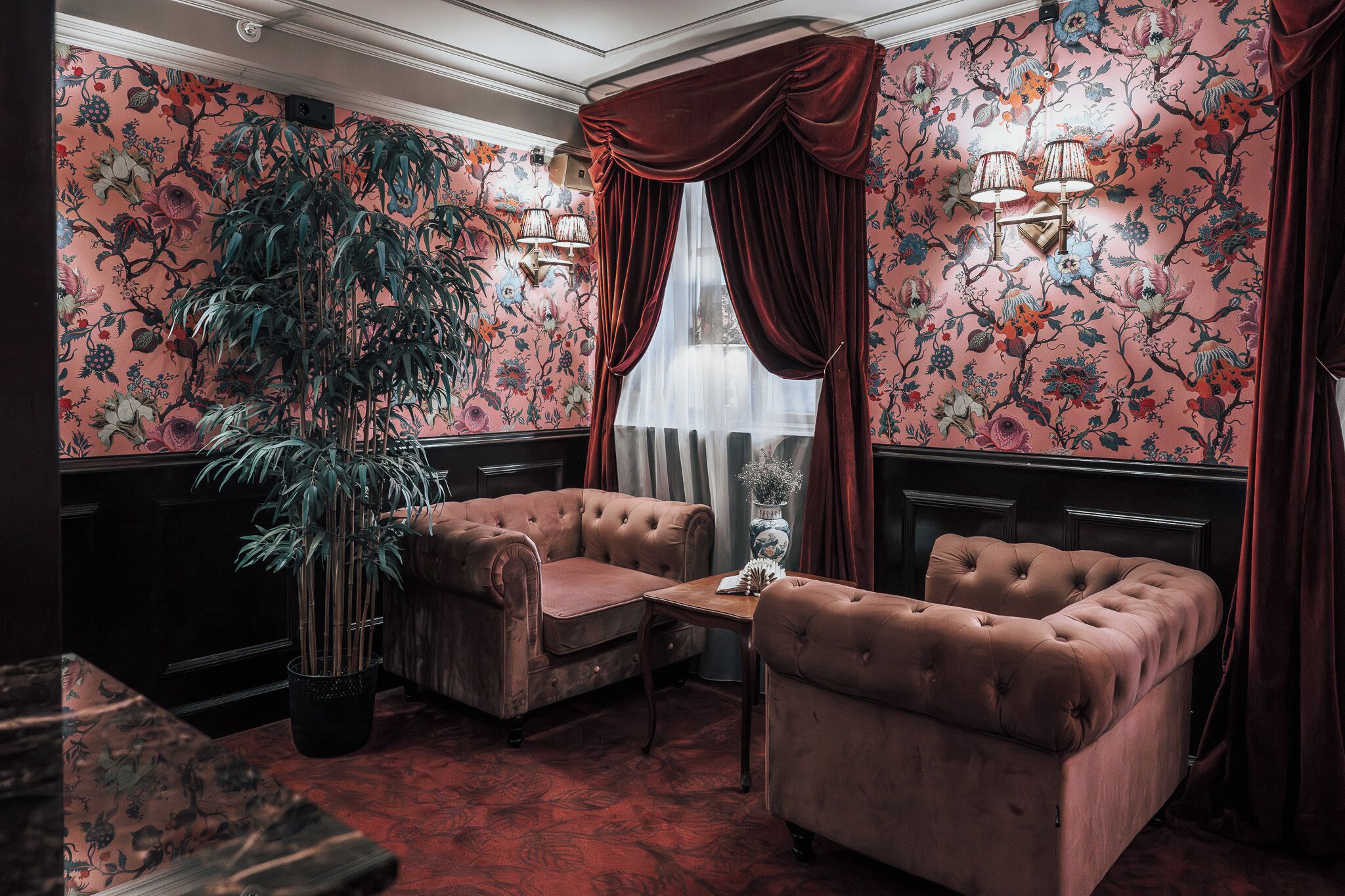 Una habitación con papel tapiz floral, una planta grande en maceta, dos sillones capitonados, una mesita y una ventana con cortinas rojas.