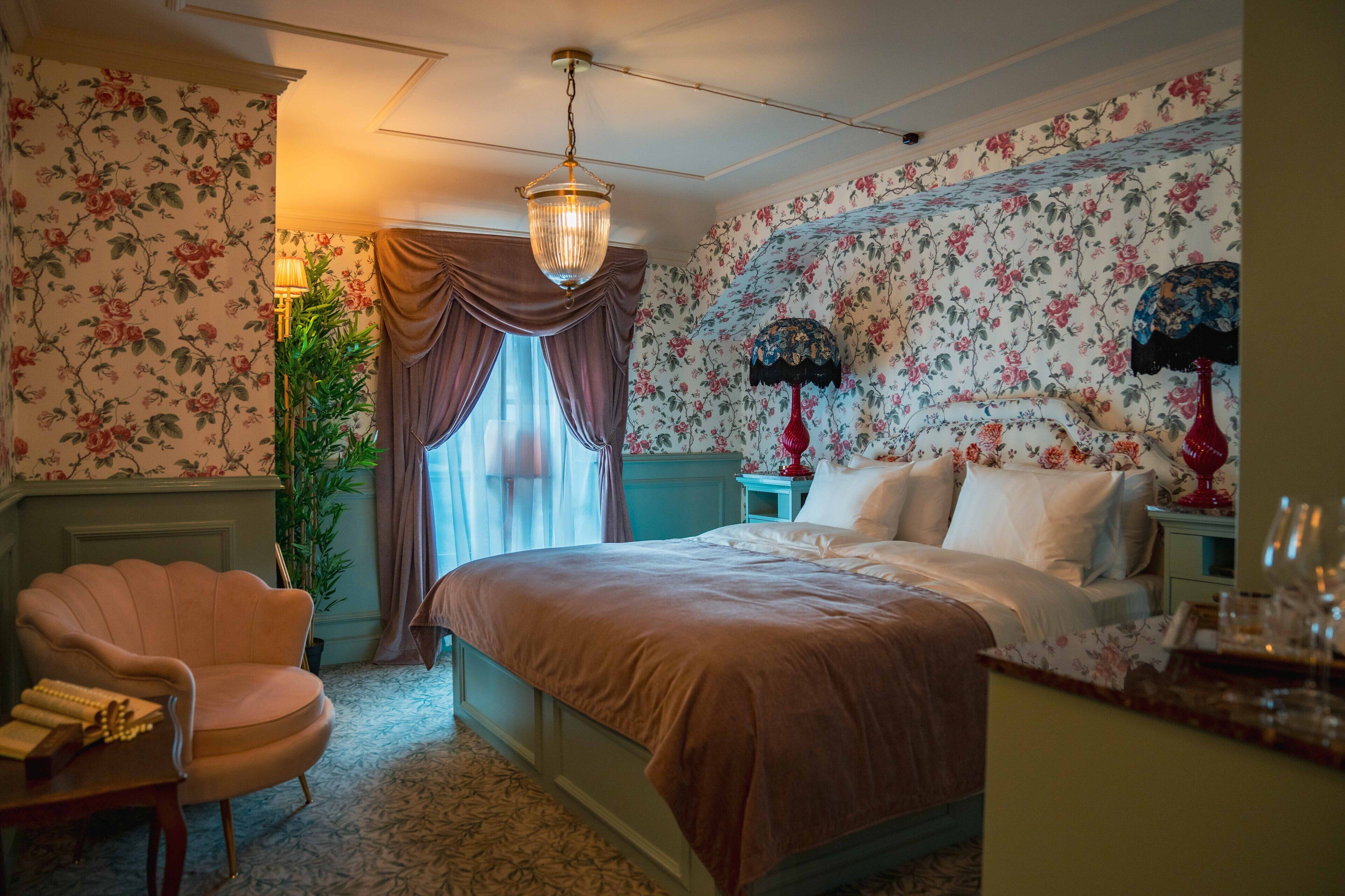 Un dormitorio con cama, una silla, una lámpara y un papel tapiz con motivos florales.
