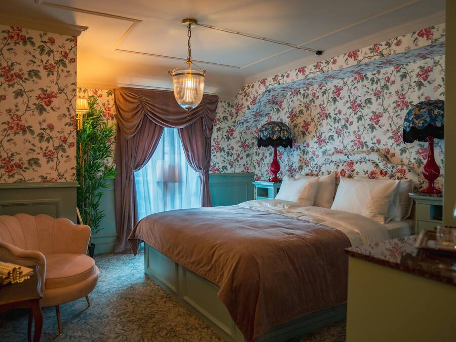 Un dormitorio con cama, una silla, una lámpara y un papel tapiz con motivos florales.