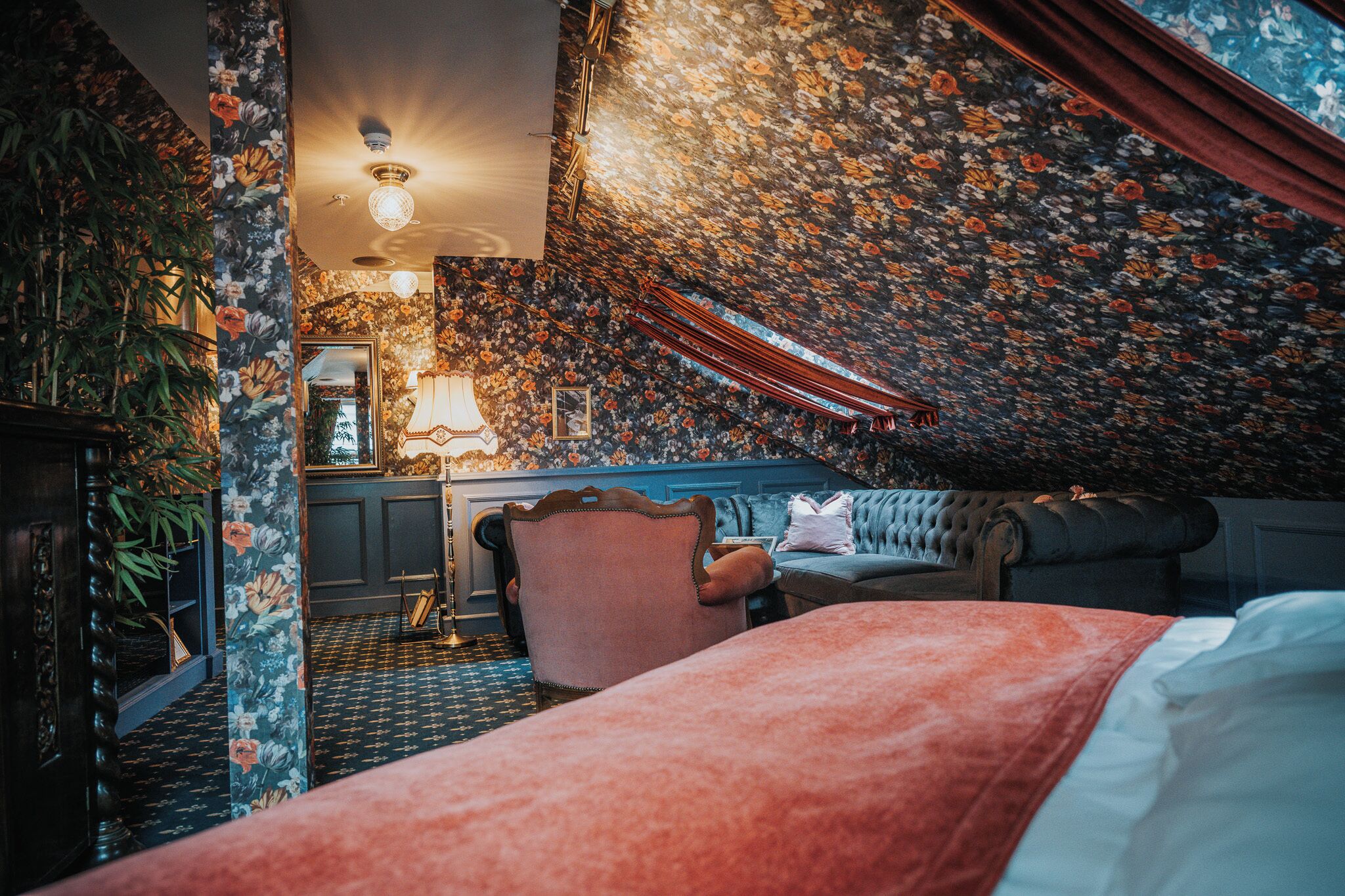 A bedroom with a floral wallpaper, a bed, a chair, and a desk with a lamp.