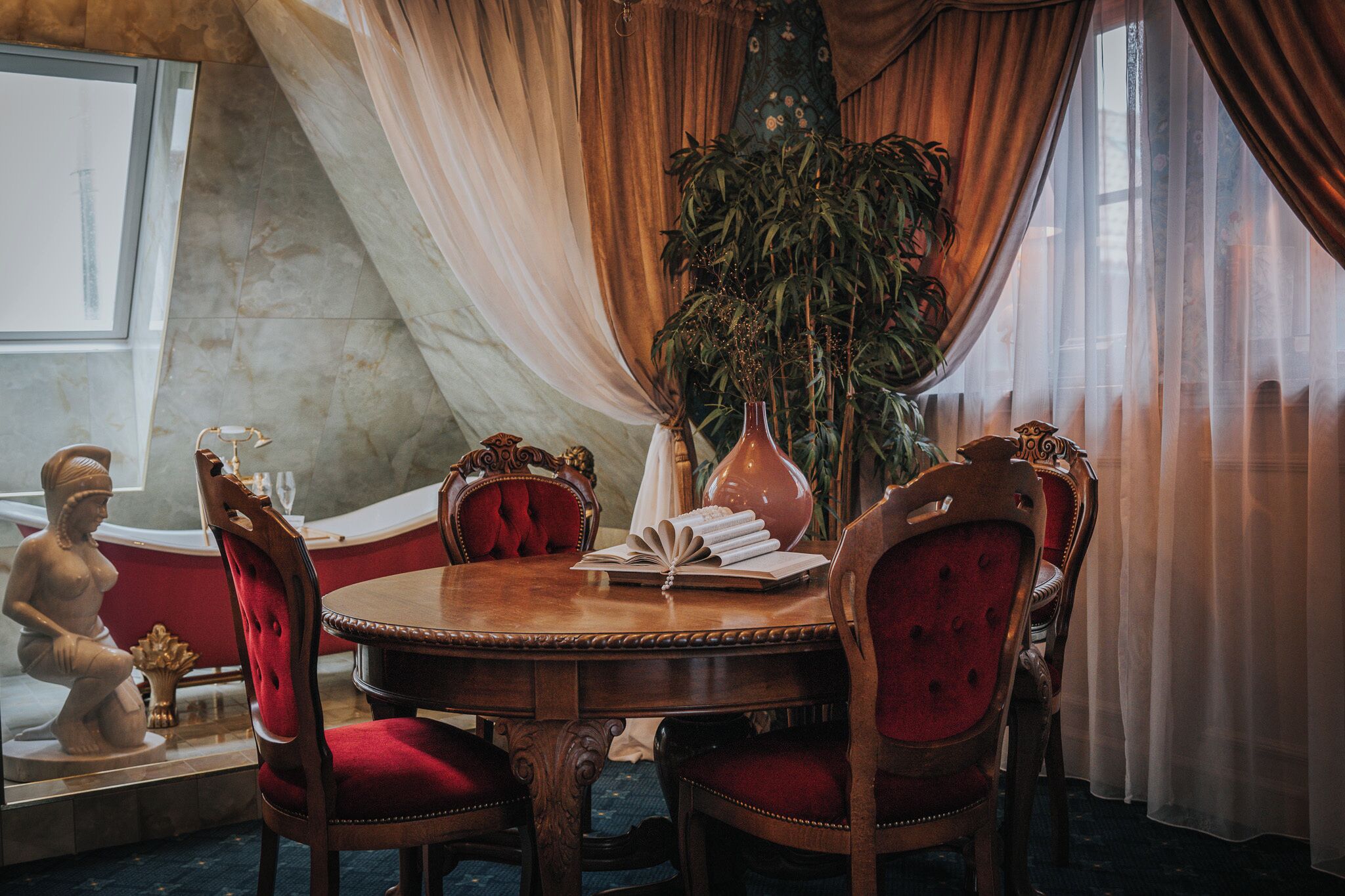 A dining area with a round wooden table, red upholstered chairs, a vase, and a potted plant.