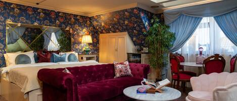 A room with a bed, a red velvet sofa, a round coffee table, and a pink armchair.