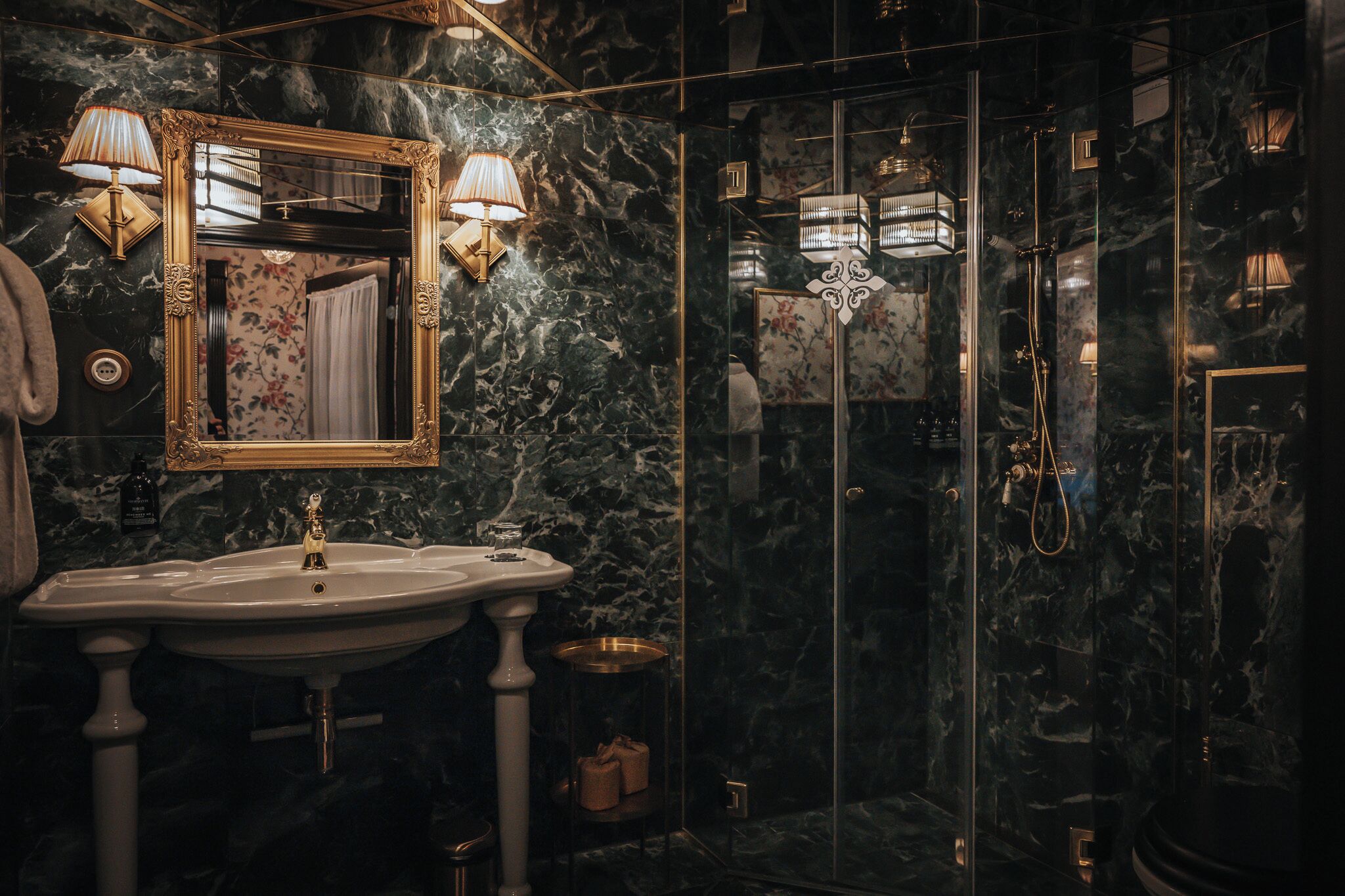 A bathroom with a marble sink, a large mirror with ornate gold frame, and a glass-enclosed shower area with a chandelier.