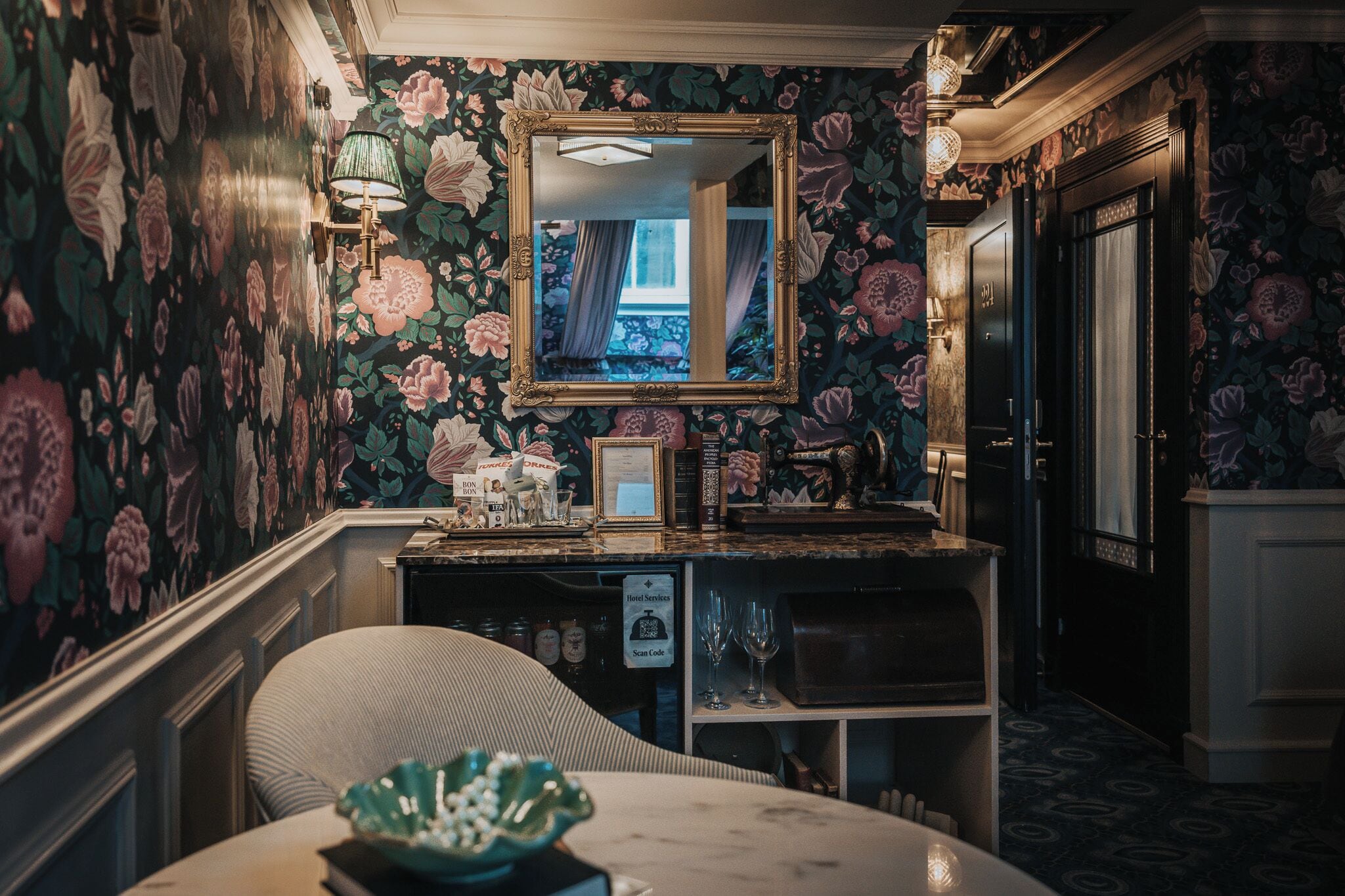Une pièce avec un papier peint à motifs floraux, un miroir, une table avec un bol, et un coin bar avec des verres et une enseigne.