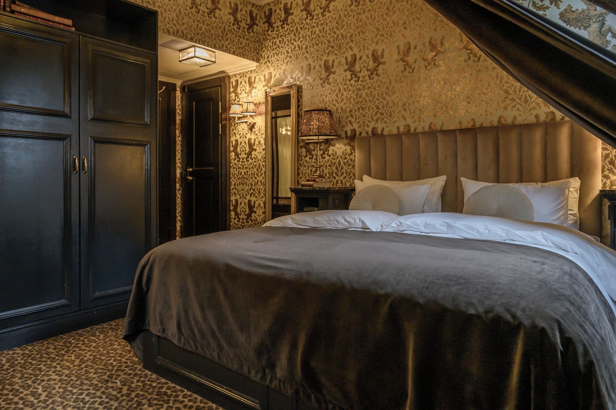 A bedroom with a large bed, dark wooden wardrobe, and patterned wallpaper.