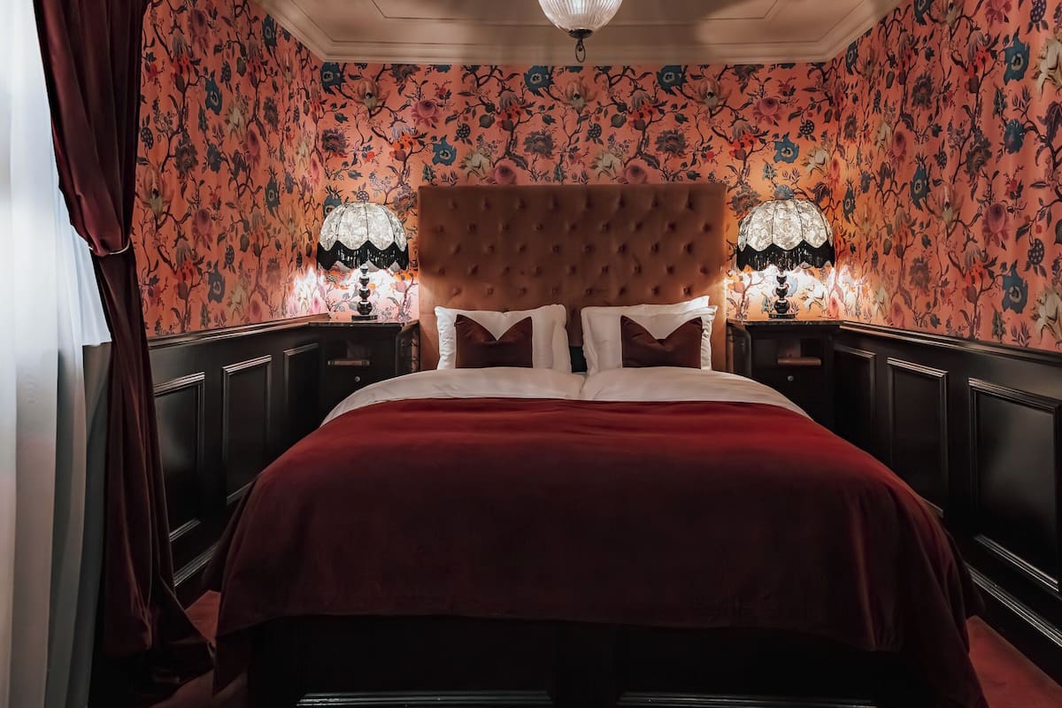 Ein Schlafzimmer mit einem gesteppten Kopfteil, floralem Wandpapier und einer roten Bettdecke.
