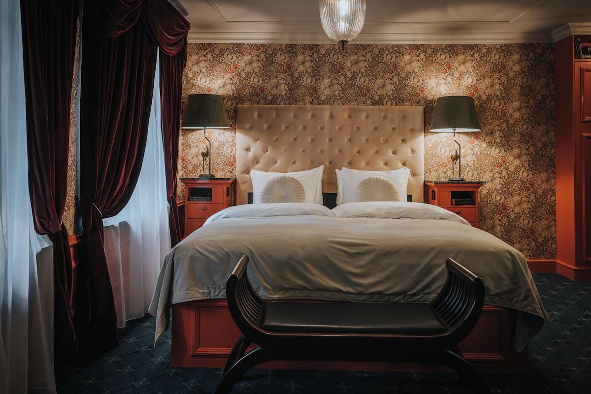 Ein Hotelzimmer mit einer gepolsterten Kopfstütze, zwei Nachttischlampen, einem Bett mit weißen Leinen und einem dunklen Holzstuhl.