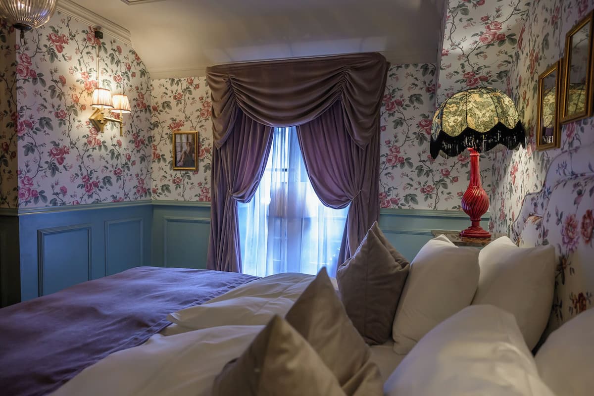 Ein Schlafzimmer mit floraler Tapete, einem Bett mit vielen Kissen, einem Fenster mit violetten Vorhängen und einer roten Lampe auf einem Nachttisch.