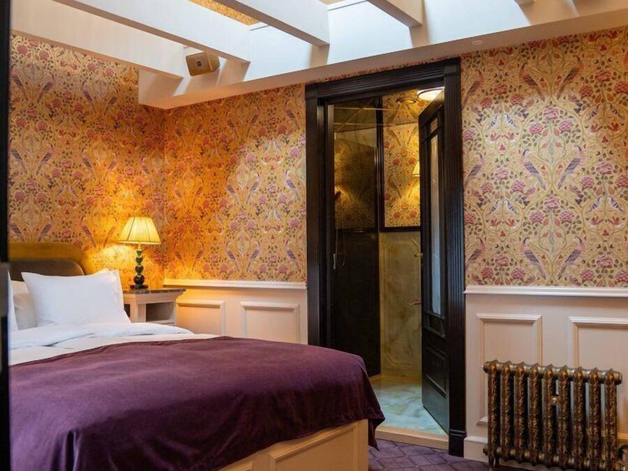 Een slaapkamer met een groot bed, een nachtkastje, een lamp, een radiator en een deur die naar een andere kamer leidt.