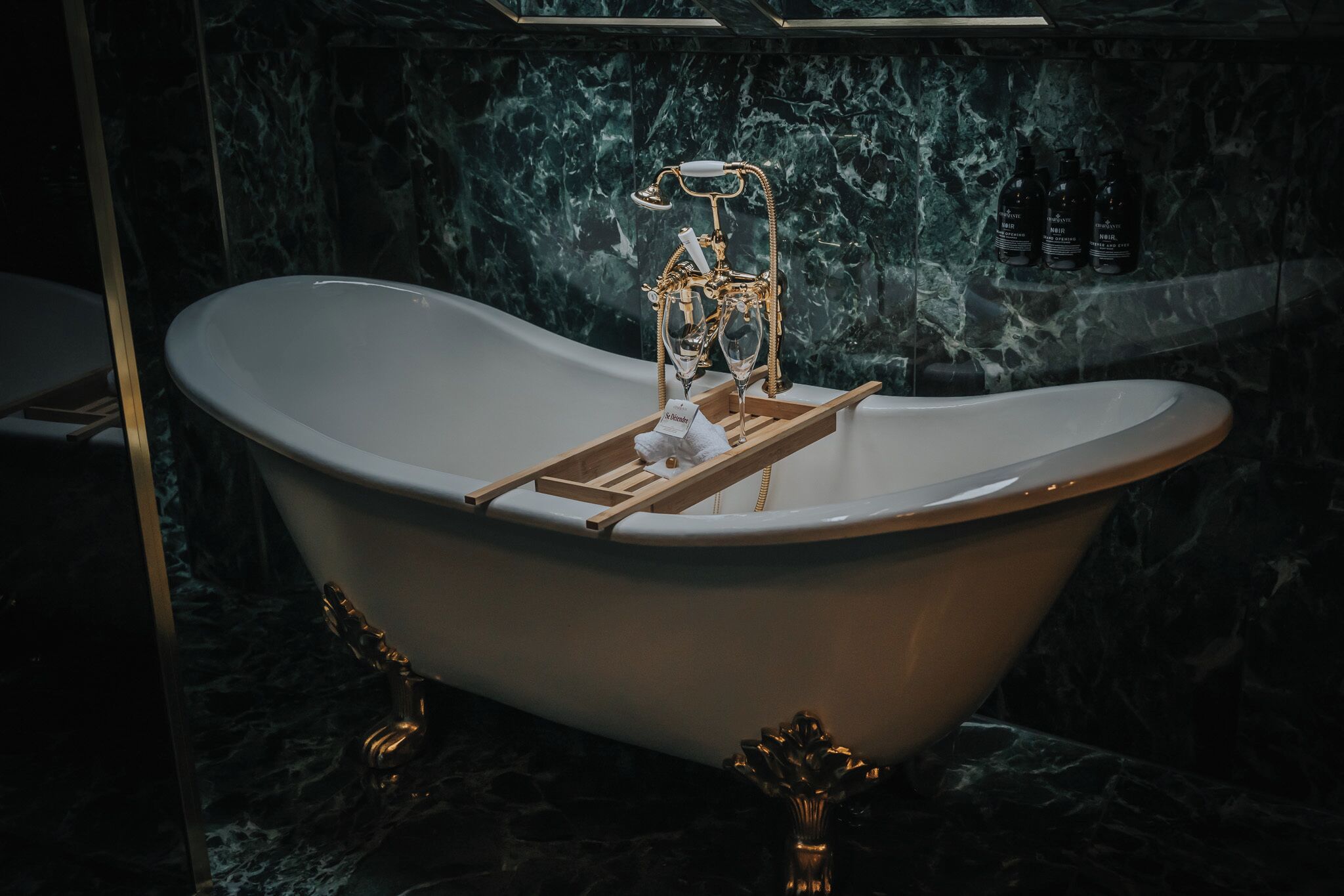 A freestanding bathtub with a wooden tray and a towel, set against a dark marble wall.