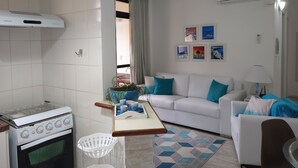 Un espacio habitable compacto con kitchenette, un sofá blanco con cojines azules, una mesa de comedor y una lámpara.