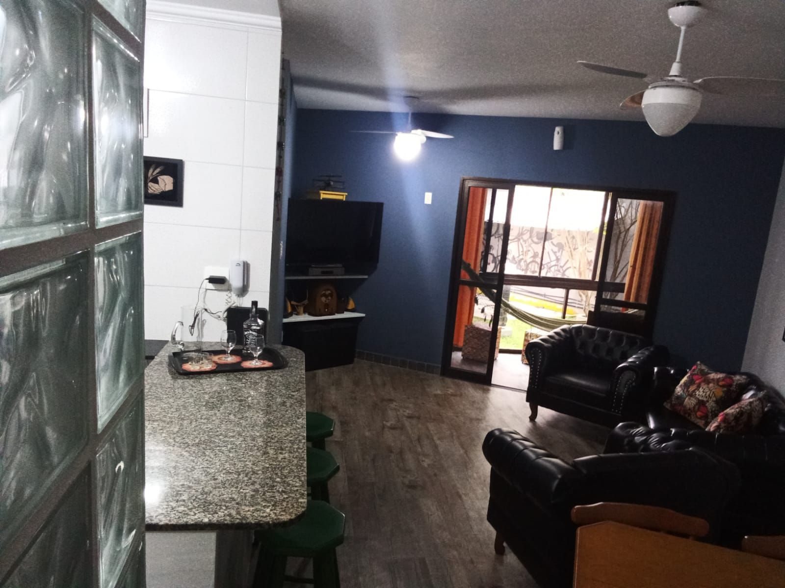 Un salon avec un canapé en cuir noir, une table basse et un téléviseur. Il y a un coin cuisine avec un comptoir en granit et des tabourets de bar. La pièce est équipée d’un ventilateur de plafond et d’une grande fenêtre avec des rideaux.