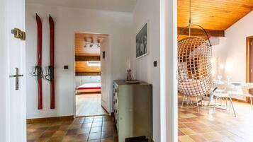 A room with a wooden ceiling, a dining area with a hanging chair, and a view into another room with a bed.
