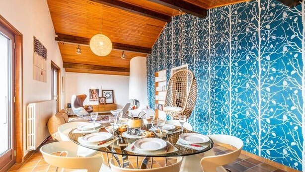Een eetgedeelte met een ronde glazen tafel, gedekt voor zes personen, omringd door moderne stoelen. Een rieten stoel staat tegen een muur met een blauw boompatroon.
