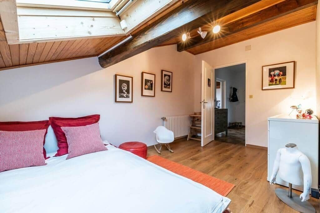 Ein Schlafzimmer mit Holzbalkendecke, einem Bett mit rot-weiß gestreifter Bettwäsche, einem weißen Sessel und einem Heizkörper.