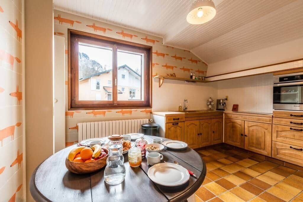 Una cocina con armarios de madera, una mesa redonda de comedor con cosas para el desayuno, y un ventanal con vistas a casas.