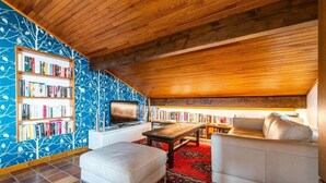 Een gezellig hoekje om te lezen, met een houten plafond, een boekenkast, een comfortabele bank en een televisie.
