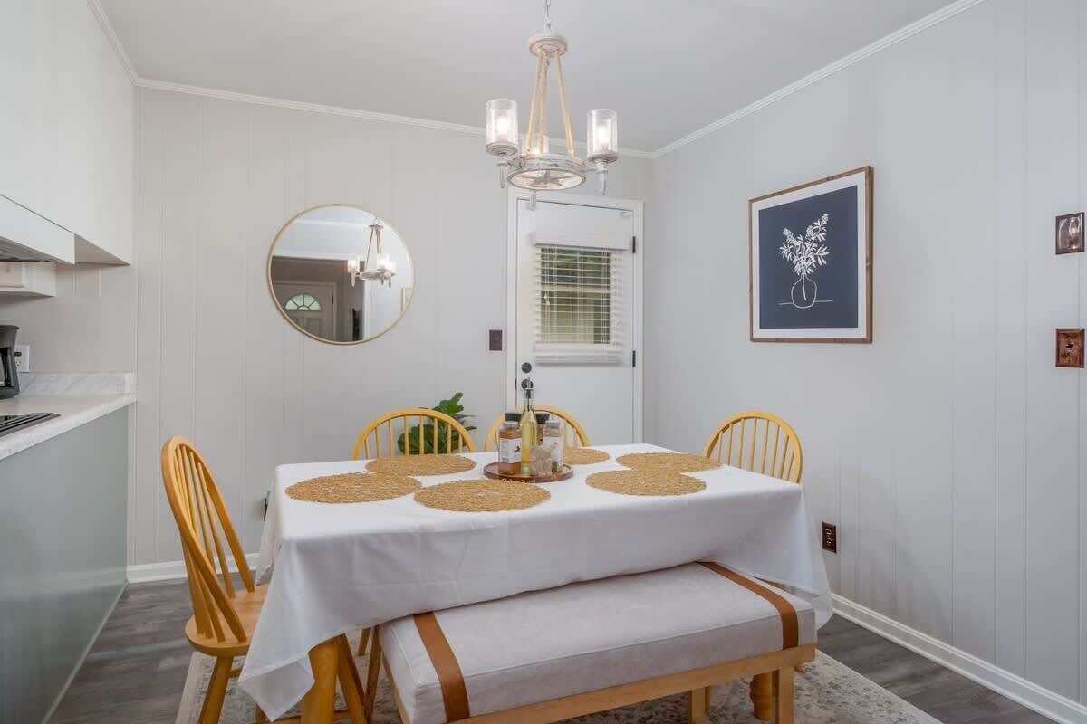 A dining area with a white table, wooden chairs, a chandelier, and a framed artwork on the wall.