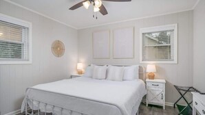 A bedroom with a white bed, two bedside tables with lamps, a ceiling fan, and a window with blinds.