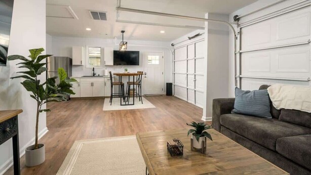 A modern living room with a wooden floor, a dining area with a table and chairs, a grey sofa, and a TV mounted on the wall.