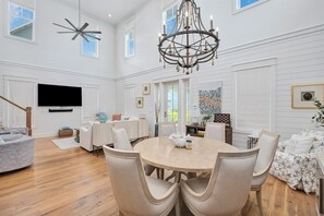A spacious living room with a round dining table, comfortable seating, a chandelier, and a large TV mounted on the wall.