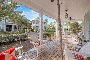 A covered porch with white chairs and a swing, overlooking a residential area with houses and greenery.