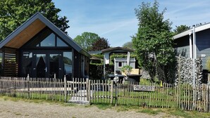 A modern building with a wooden facade and large windows, surrounded by a gravel area and a wooden fence. - Tiny House Holland by the lake. A quiet, comfortable break on the beach by the water. (Lathum)