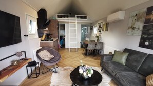 A spacious living area with a sofa, a round coffee table, and a dining area with chairs and a table. - Tiny House Holland by the lake. A quiet, comfortable break on the beach by the water. (Lathum)