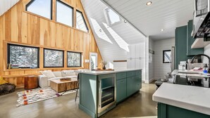 Una cocina moderna con horno empotrado, un comedor con mesa y sillas blancas, y ventanas grandes que ofrecen vistas a árboles cubiertos de nieve.