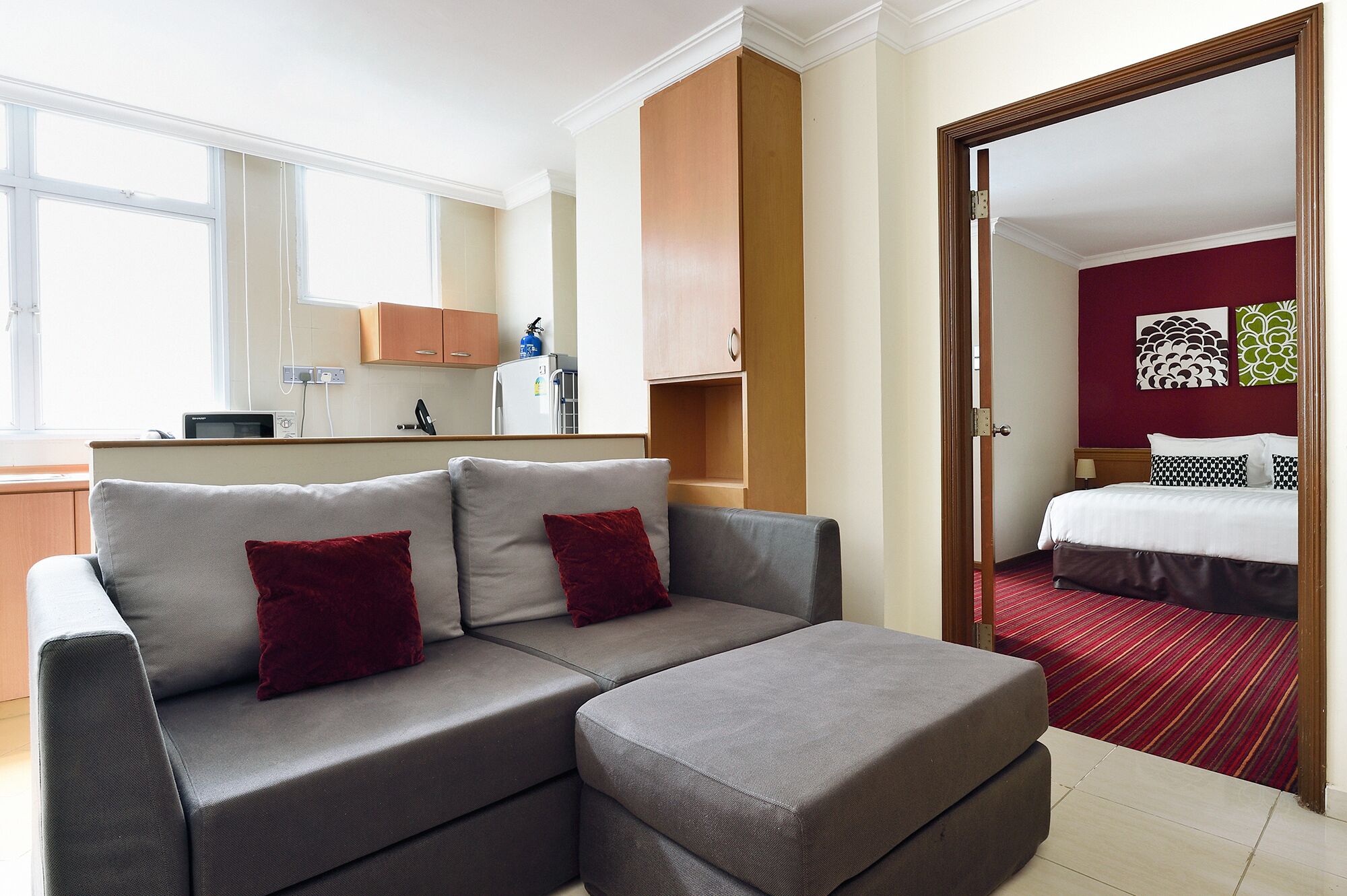 Een moderne hotelkamer met een grijze bank, rode kussens, een kleine kitchenette, een bed met een rood-zwarte gestreepte dekbedovertrek en een spiegel die de kamer weerspiegelt.