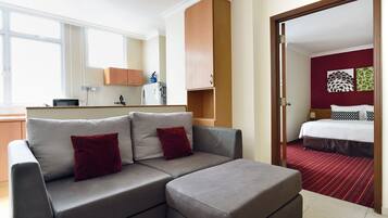 Een moderne hotelkamer met een grijze bank, rode kussens, een kleine kitchenette, een bed met een rood-zwarte gestreepte dekbedovertrek en een spiegel die de kamer weerspiegelt.