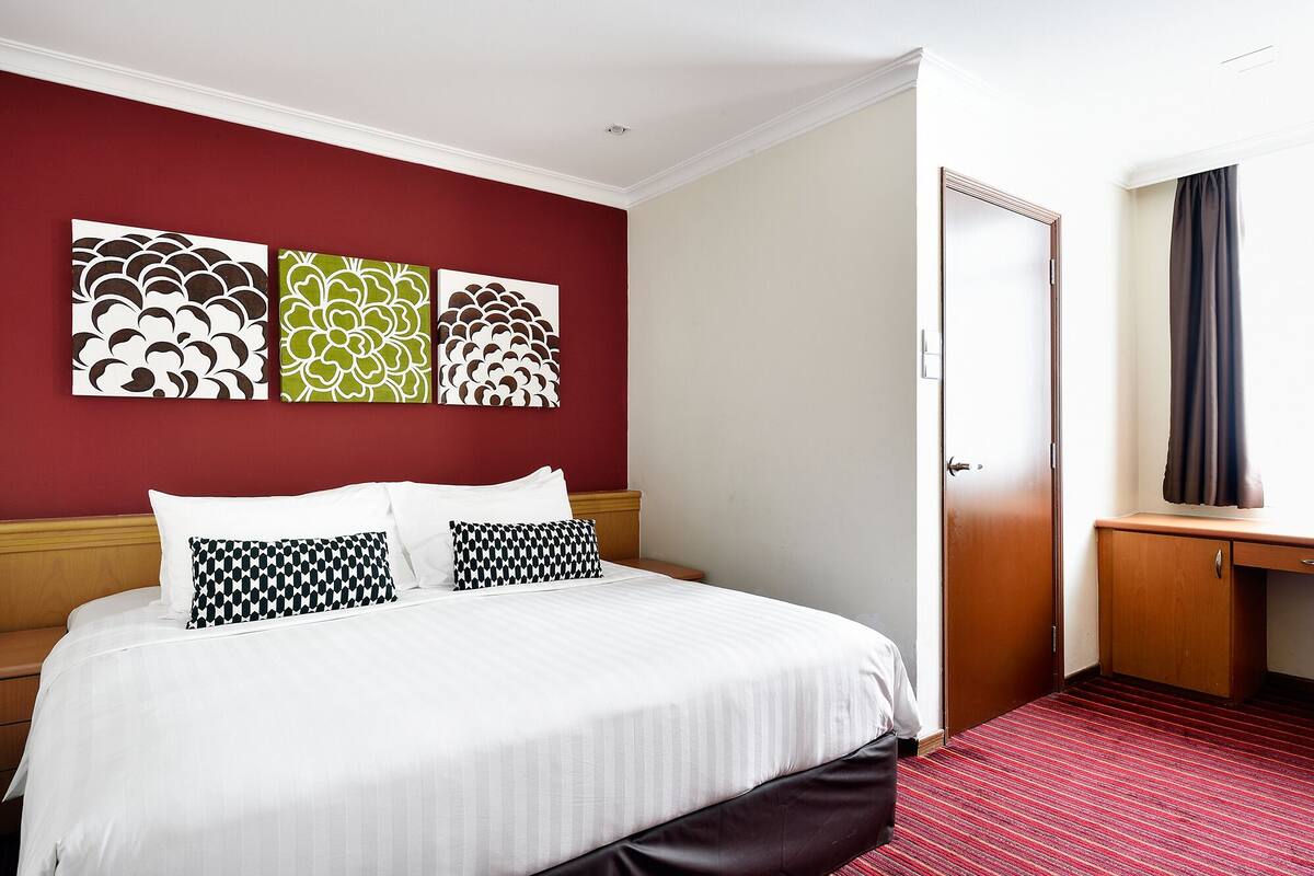 A hotel room with a bed, a wooden nightstand, a desk, and a door.