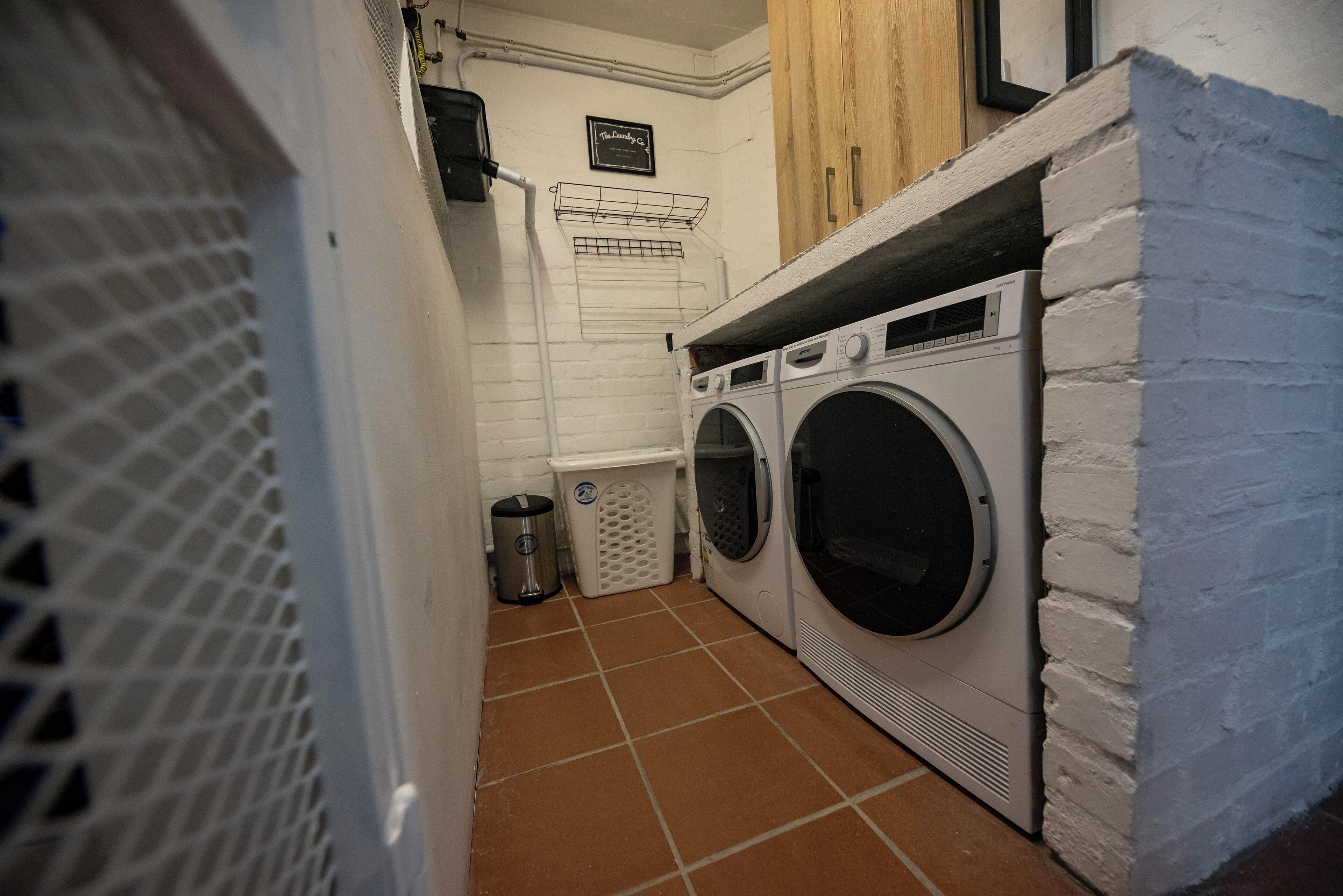 A laundry room with two front-loading washing machines, a drying rack, and a trash can.
