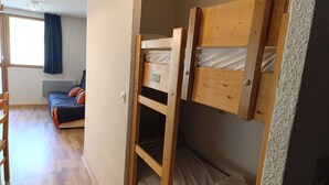 En våningssäng med en träram och en madrass, en trästol, ett litet bord och en soffa i ett rum med trägolv och ett fönster med gardiner.