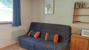 A room with a sofa, a radiator, a window with curtains, a framed picture, and a shelf with books and pens. - Paradiski closed to the slopes and shops vallandry (LANDRY)
