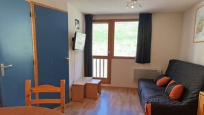 Ett rum med trägolv, en svart soffa med orange kuddar, en trästol, ett litet träbord och ett fönster med gardiner.