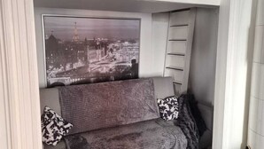 Una habitación con una cama litera, un cartel de paisaje urbano en la pared, un sofá gris con cojines estampados y una manta roja en el suelo.
