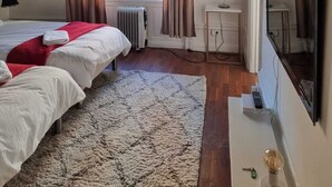 Habitación de hotel con dos camas, un radiador, un escritorio con lámpara, un televisor y obras de arte en las paredes.