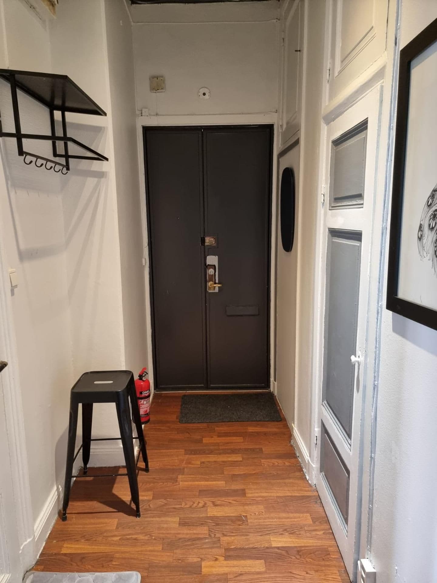 A hallway with a wooden floor, a black door with a keyhole, a small stool, and a fire extinguisher.
