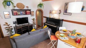 Ein Wohnzimmer mit grauem Sofa, Fernseher, Klavier und Esstisch mit Obstschale.