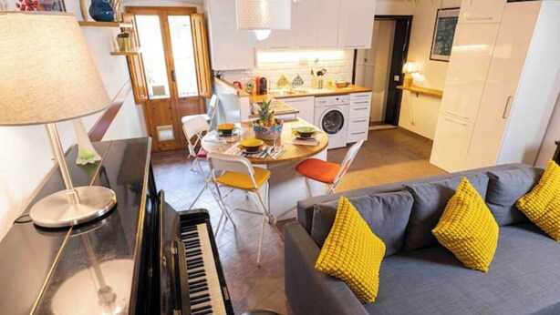 Ein modernes Wohnzimmer mit einem Klavier, einem grauen Sofa mit gelben Kissen und einem Essbereich mit Tisch und Stühlen.