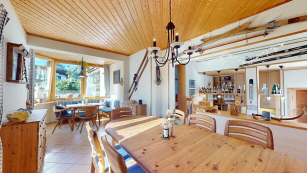 Une table à manger en bois, des chaises, un lustre et un coin cuisine avec des étagères et des ustensiles.